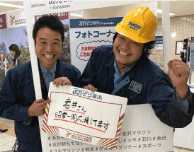 金沢マラソンのフォトコーナーで、ヘルメット姿の社員と応援メッセージボードを持つ2人